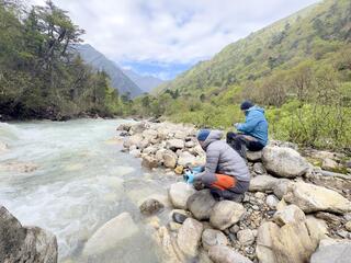 Two people sit on the edge of a mountain river collecting samples to test for DNA