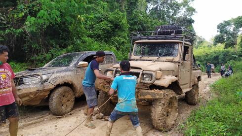 Two men in blue shirts tow a muddy Jeep along a muddy forest path
