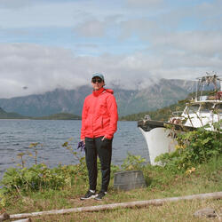 A woman in a red coat stands outside with harbor and mountains in background