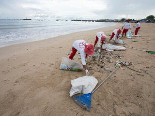 People in white shirts and red hats pick up plastic waste on a beach in Indonesia.