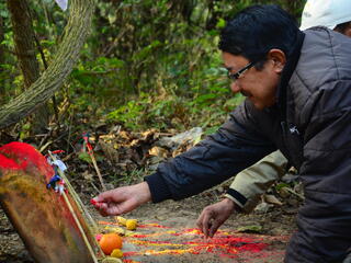 A man kneels down on the ground in the forest to place red powder on a stone