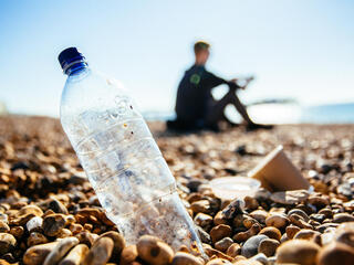 A plastic bottle on a pebble beach sits in the foreground with a sillouhette of a man in the background