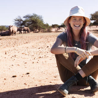Behati sits on the ground smiling at the camera with rhinos in the far background