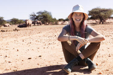 Behati sits on the ground smiling at the camera with rhinos in the far background