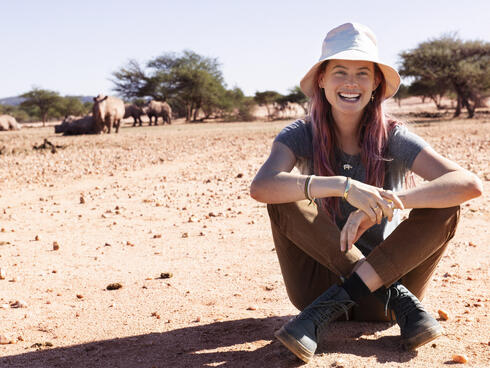 Behati sits on the ground smiling at the camera with rhinos in the far background