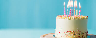 A white birthday cake with rainbow sprinkles and several lit candles sits on a tabletop 