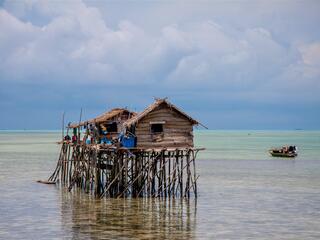 Small woven houses sitting on top of a system of stilts out in clear blue water with a green boat anchored nearby