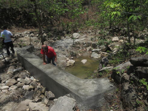 A man in a red shirt holds a rock and places a hand on a human-made dam for an artificial pond