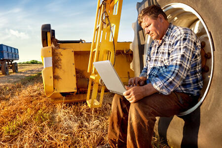 Man sits outside with farm equipment using laptop