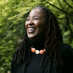 Smiling headshot of Camille Dungy. Her locs fall below the shoulder, and she is wearing all black, a large red and gold necklace. She looks off to the right. In the background are trees.