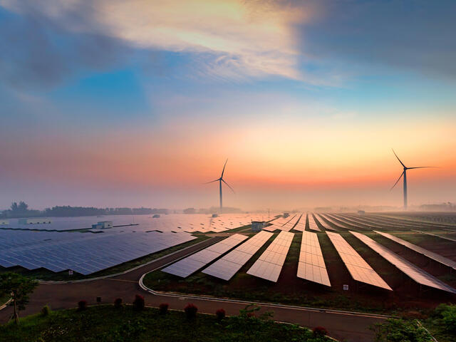Landscape photo of a field of solar panels at sunrise. There are two wind turbines in the distance. Everything is shrouded in a light mist.