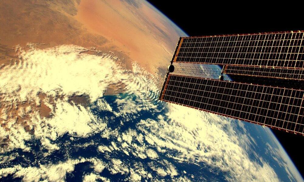 An image of Earth, taken from space that shows a desert climate on earth and has a satellites solar array in the foreground