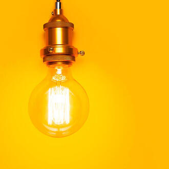 A round Edison bulb with brass hardware hangs from a black cord and is lit up. It is in front of a warm yellow background