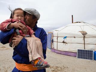 A father holds his young daughter in his arms and kisses her on the cheek in front of a yurt