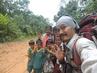 Man with red backpack stands on a forest path with group of children all waving at the camera