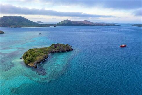 A chain of islands surrounded by barrier reefs in blue water with a red boat anchored offshore