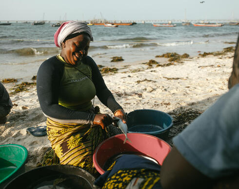 A woman seated outside on a beach cleans a fish