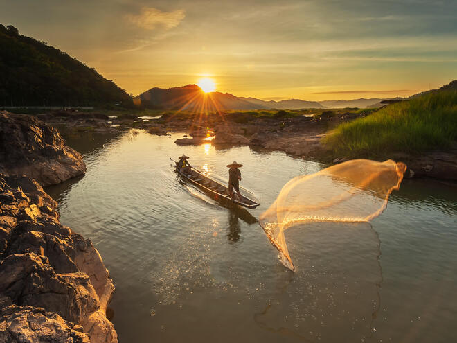 Two people cast a fishing net into the Mekong River as the sun rises