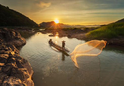 Two people cast a fishing net into the Mekong River as the sun rises
