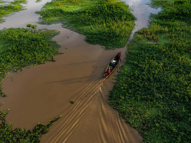 Two people drive a small boat through muddy waters and green marshland 