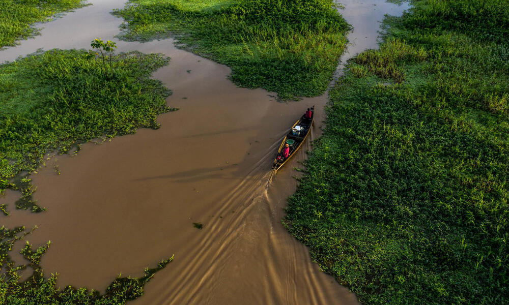 Two people drive a small boat through muddy waters and green marshland 