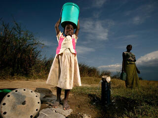 A girl in a white dress holds a green bucket above her head and smiles at the camera