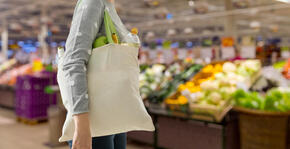 A woman with a beige tote bag walks through the produce section of a grocery store