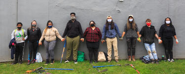 A group of people stands against a wall holding hands; most are wearing masks and there are gardening tools on the grass in from of them