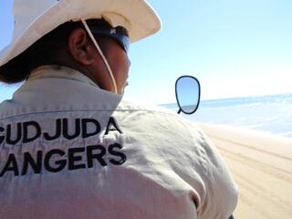 View of a person's back against the beach. Wearing a khaki hat and jacket that says "Gudjuda Rangers"