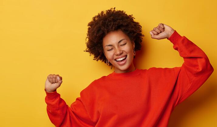 Woman celebrates with her eyes closed and her hands up against a solid background