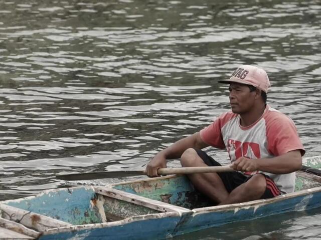 A man in a tshirt and pink baseball cap sits inside a skinny blue canoe on the water