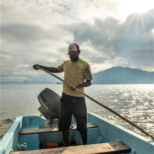 A man in a yellow tshirt and baseball cap stands in a small blue fishing boat holding a long paddle out at sea with the sun shining and a mountain range behind him