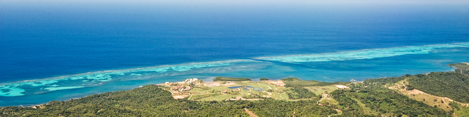 Aerial view of a coastline in Roatan, Bay Islands, Honduras. The ocean is a deep blue. At the barrier reef, the water is turquoise.