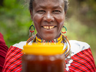 A woman in colorful clothing smiles and holds out a jar of honey