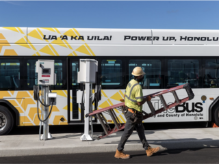 A worker carrying a ladder walks past an electric bus and charging station.