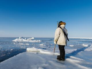 A person wearing full Arctic gear holds a harpoon, standing on the sea ice and looking out into the frozen ocean.