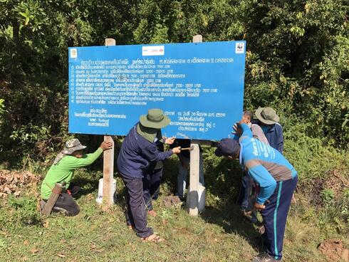 A group of people install a large blue sign along the edge of a wooded area