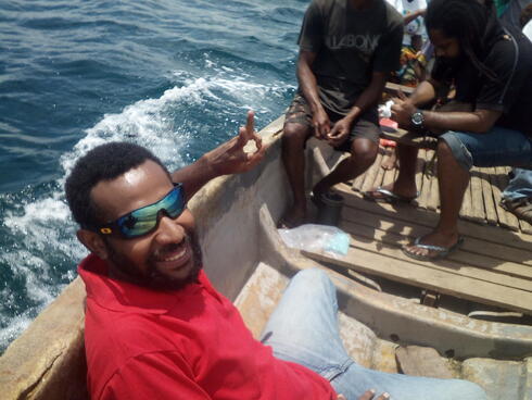 A man wearing sunglasses and a red shirt smiles at the camera while sitting in a small wooden boat on the ocean