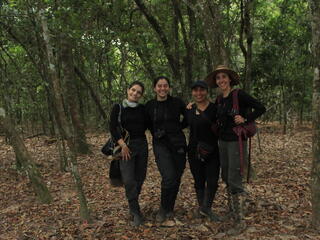 Four women and scientists of color stand together embracing shoulders and smiling at the camera. They are under a canopy of trees.
