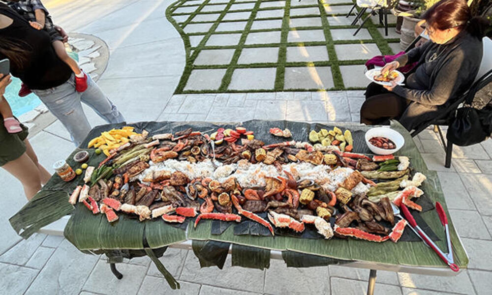 People gather around a large table covered with grilled banana leaves and food