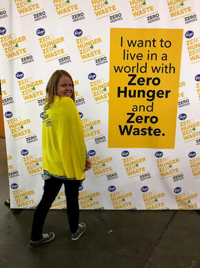 A woman stands in front of a "Zero Hunger | Zero Waste" sign