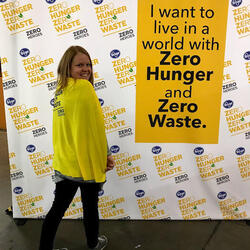 A woman stands in front of a "Zero Hunger | Zero Waste" sign