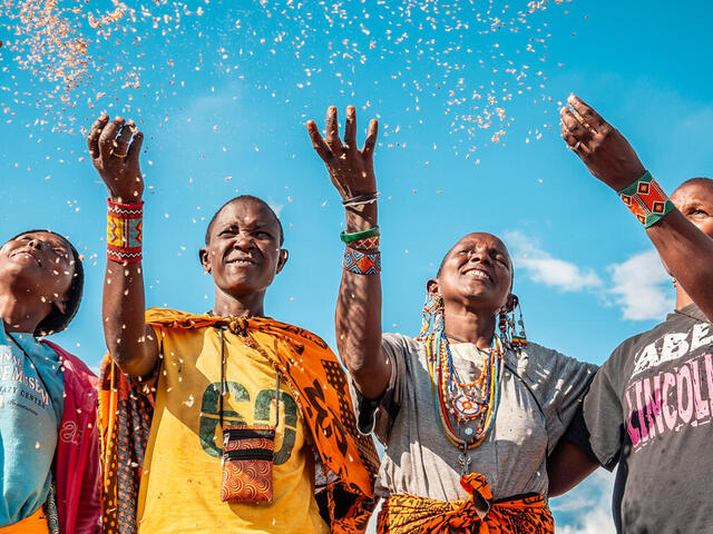Four people in colorful clothing smiling and tossing seeds into the air