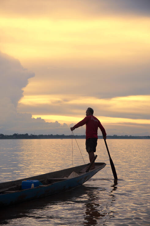 A fisherman stands on the tip of his canoe out on the river preparing to throw his net onto the water