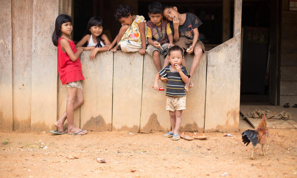 A group of small children sit along a wooden wall