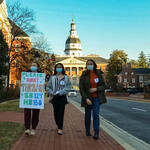 Three individuals walk down the brick street in Annapolis. One youth is holding a sign that reads 'Please don't throw out SB124 HB150.'