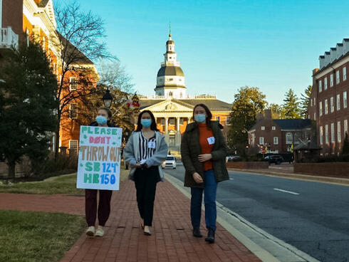 Three individuals walk down the brick street in Annapolis. One youth is holding a sign that reads 'Please don't throw out SB124 HB150".