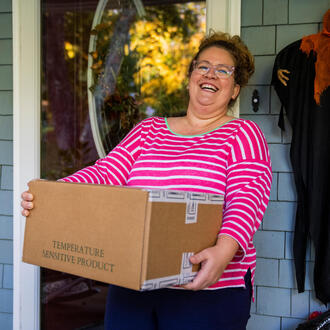A woman in a pink shirt smiles on a porch holding a box of vegetables to mail