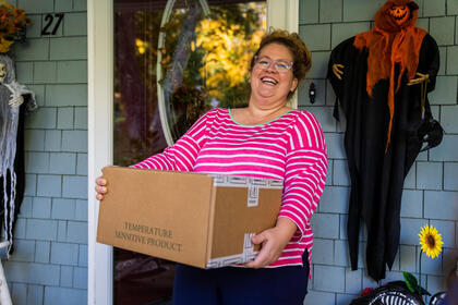 A woman in a pink shirt smiles on a porch holding a box of vegetables to mail