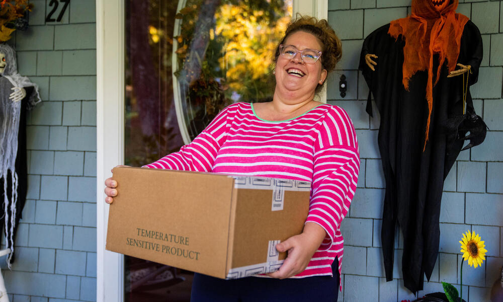 A woman in a pink shirt smiles on a porch holding a box of vegetables to mail